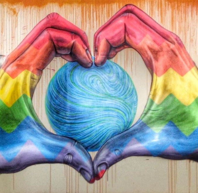 Mural on a wall in rainbow colors depicting a large sphere in blue and green. The sphere is held by two hands, grasped to display the outline of a heart with the fingers.