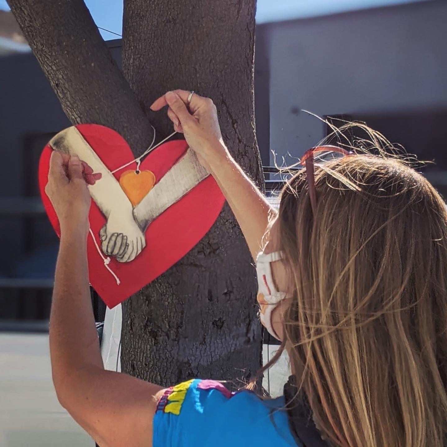 Young girl with blond hair hanging a wood carved heart on a tree. Wood carved heart depicts two hands grasping each other.