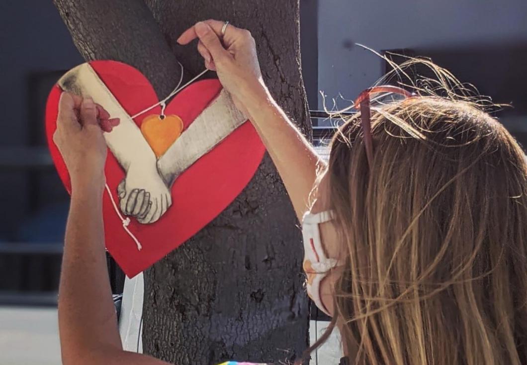 Young girl with blond hair hanging a wood carved heart on a tree. Wood carved heart depicts two hands grasping each other.