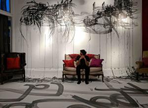 man siting on red couch in white room with black inked Persian-Arabic calligraphic scripts on the walls and floor