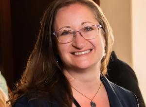 A woman with light skin, brown hair, and glasses smiles.