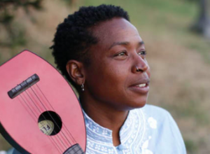 Headshot of poet Sevan Kelee Boult. She is a Black queer woman in a white shirt and holder a small pink guitar.